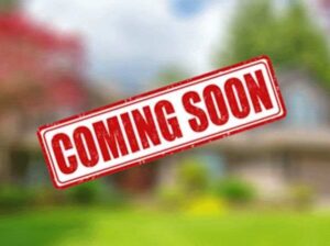 Red and white stamp with the text "COMING SOON" on it. The background is a blurry image of a house.